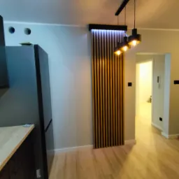 Nowoczesne wnętrze mieszkania z drewnianymi lamelami podświetlonymi ledami, wiszącą lampą z trzema kloszami i widokiem na kolejne pomieszczenie, ukazujące jasną podłogę.