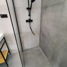 Nowoczesna kabina prysznicowa walk-in z odpływem liniowym, czarną armaturą i szarymi płytkami imitującymi beton.