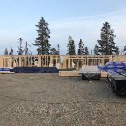 Daniel Rosiek BUDOWNICTWO - Surowa drewniana konstrukcja budynku w trakcie budowy, widoczne bale i słupy, na pierwszym planie materiały budowlane i przyczepka, w tle drzewa i niebo o zmierzchu.