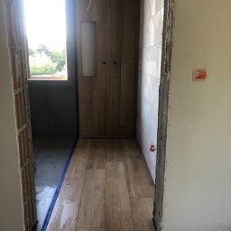 Daniel Rosiek BUDOWNICTWO - Wnętrze łazienki w trakcie prac wykończeniowych: ułożona podłoga z płytek imitujących drewno, częściowo wykończona ściana z białych płytek o geometrycznym wzorze, widoczne zabezpieczenia taśmą...