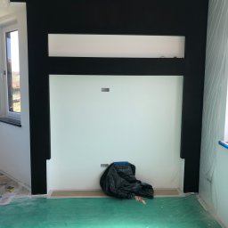 Daniel Rosiek BUDOWNICTWO - Ściana w trakcie malowania z czarną obudową, zabezpieczona folią, na podłodze zielona folia ochronna i czarny worek na śmieci.