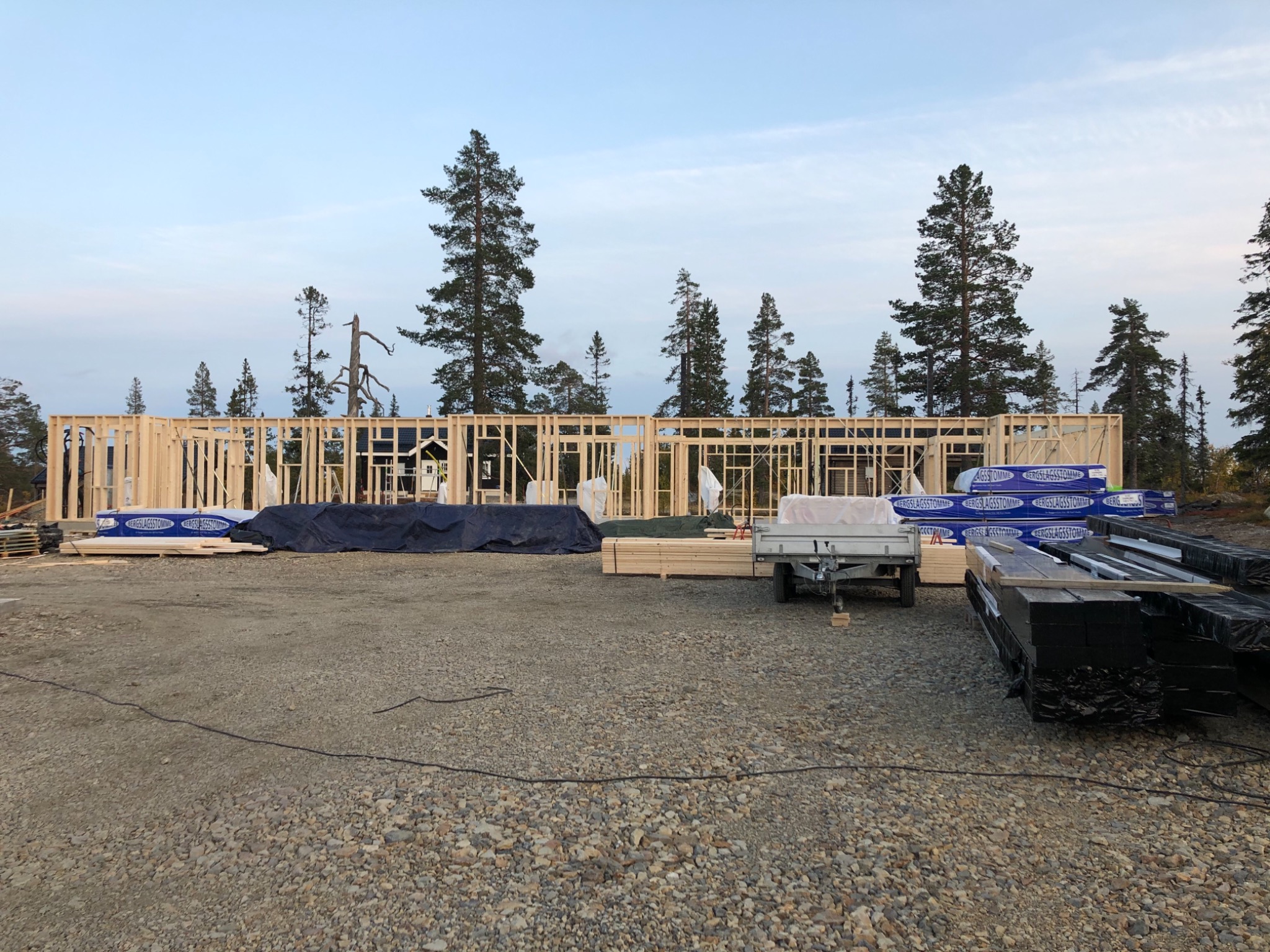 Surowa drewniana konstrukcja budynku w trakcie budowy, widoczne bale i słupy, na pierwszym planie materiały budowlane i przyczepka, w tle drzewa i niebo o zmierzchu.