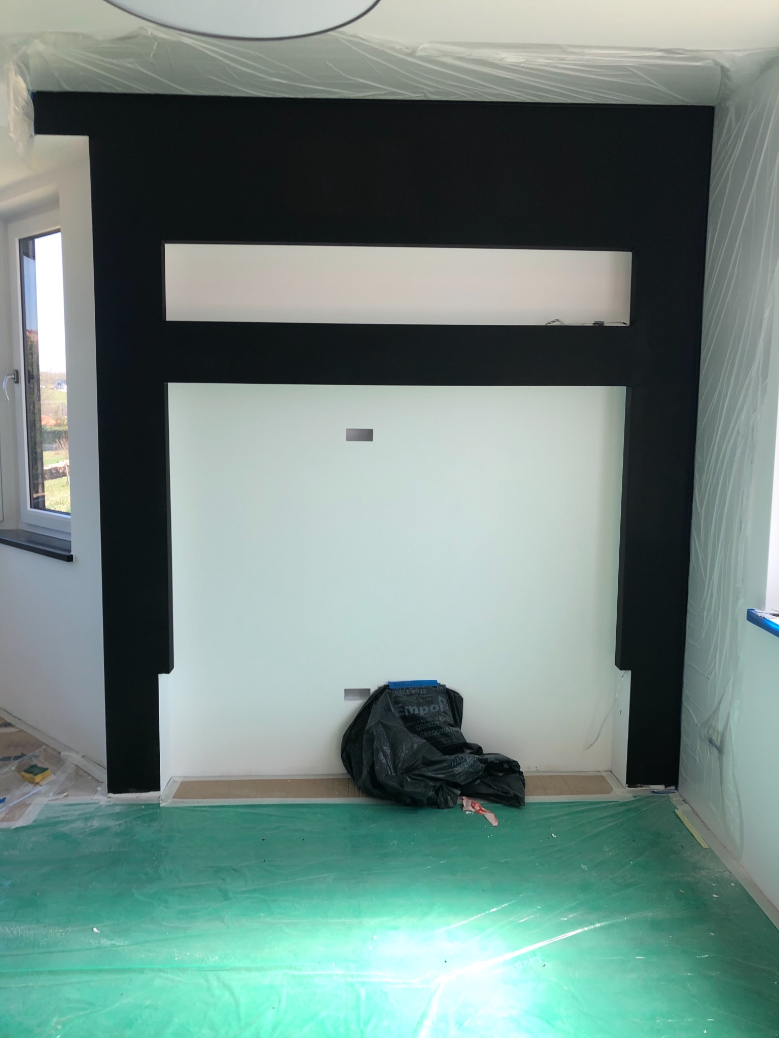 Ściana w trakcie malowania z czarną obudową, zabezpieczona folią, na podłodze zielona folia ochronna i czarny worek na śmieci.