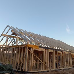 PSDom Paweł Siwek - Konstrukcja domu szkieletowego w trakcie budowy, widoczne drewniane elementy ścian i dachu na tle błękitnego nieba. Żółta folia ochronna i maszyna budowlana w tle.