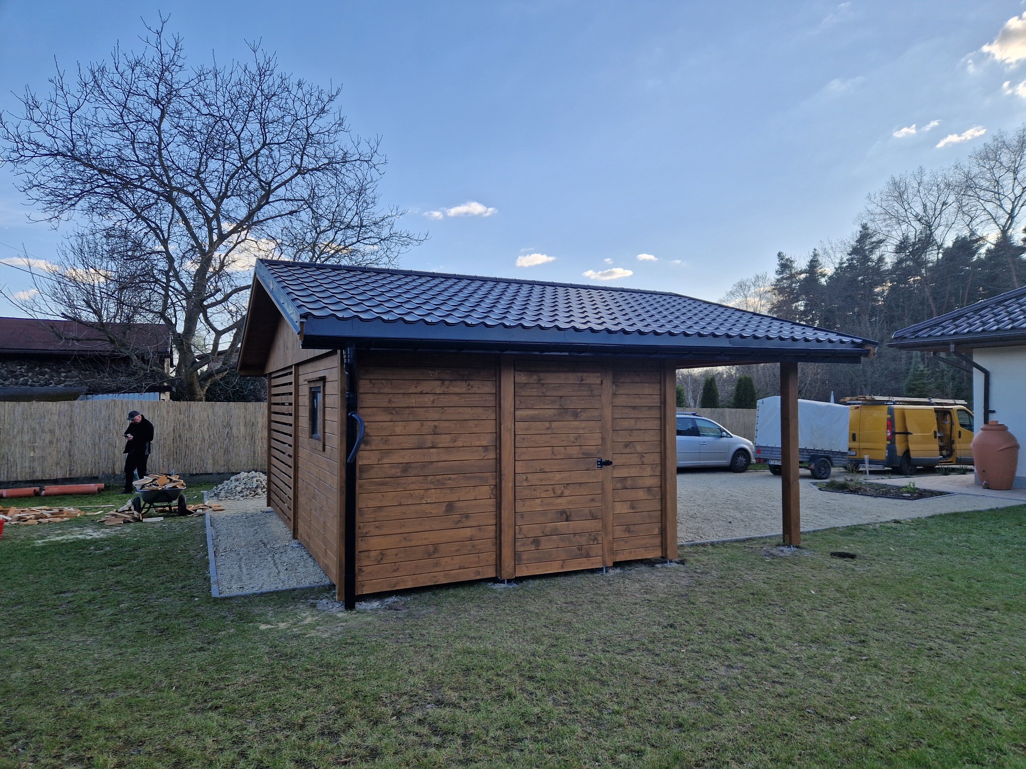 Drewniana altana z miejscem postojowym i składzikiem ogrodowym, wykończona ciemnym drewnem, z czarnym dachem, na tle zielonego trawnika i błękitnego nieba.