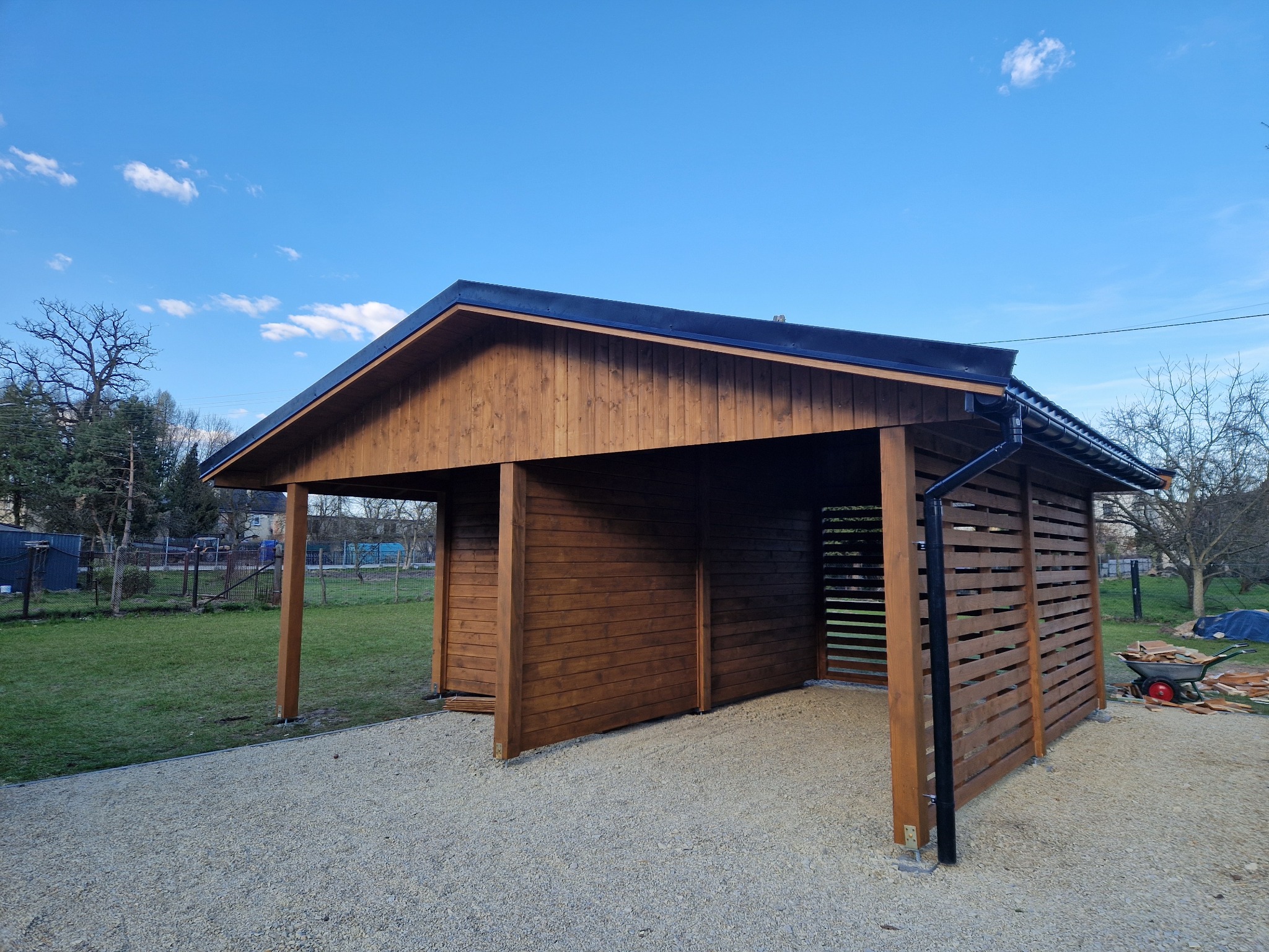 Drewniana altana z miejscem postojowym i składzikiem ogrodowym, wykończona na brązowo, z czarnym dachem i rynnami. Widok z zewnątrz, na tle błękitnego nieba.