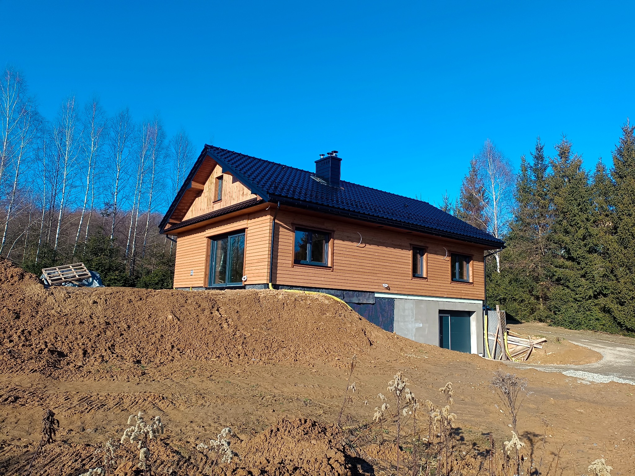 Gotowy dom szkieletowy z drewnianą elewacją i czarnym dachem, na tle błękitnego nieba i drzew. Widoczny fragment terenu z ziemią i roślinnością.