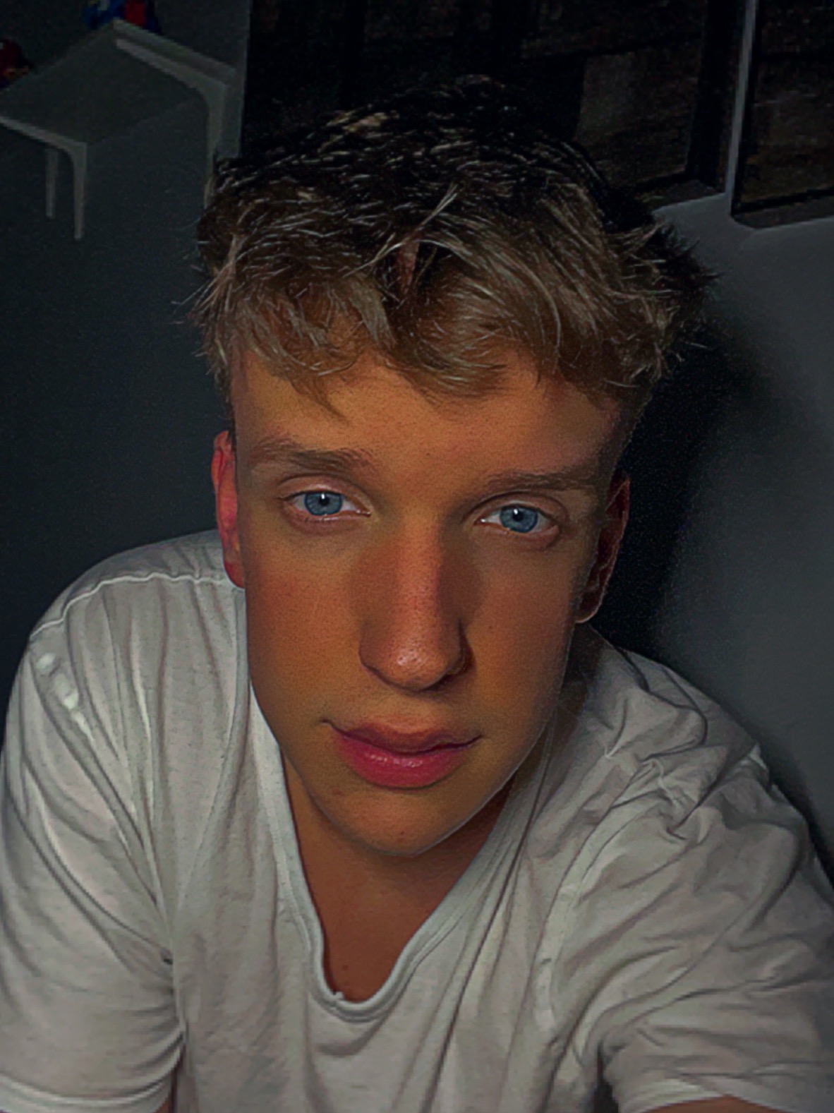 Portret młodego mężczyzny z jasnymi włosami i intensywnie niebieskimi oczami, ubrany w białą koszulkę, w ciemnym otoczeniu.