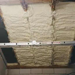 Poddasze ocieplone pianką natryskową, widoczna drabina aluminiowa z rozłożonym stabilizatorem, fragmenty krokwi i czarnej membrany dachowej.