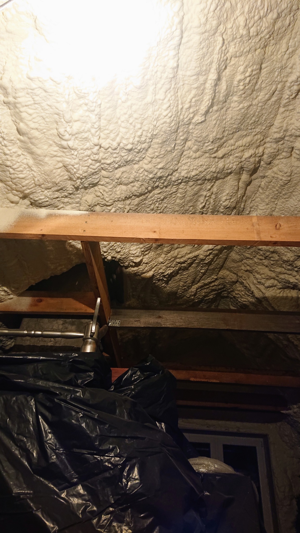 Poddasze z drewnianą konstrukcją pokryte grubą warstwą jasnej, nierównej izolacji natryskowej, zabezpieczone czarną folią przed oknem.