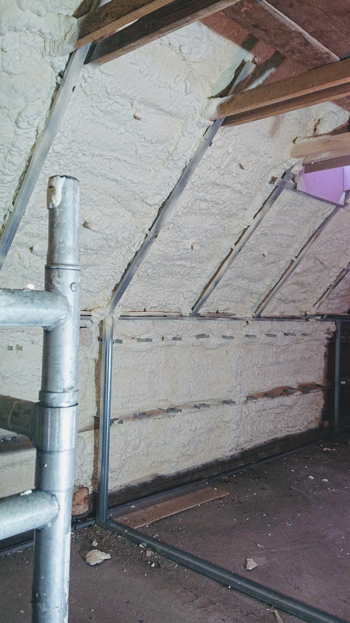 Poddasze z ukośnymi ścianami pokryte sztywną pianą izolacyjną, widoczne metalowe profile konstrukcyjne i fragment rusztowania.