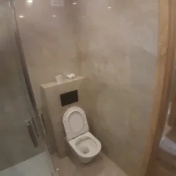 Nowoczesna łazienka z beżowymi płytkami imitującymi marmur, wisząca toaleta z otwartą klapą, kabina prysznicowa z minimalistycznymi uchwytami, oświetlenie sufitowe.