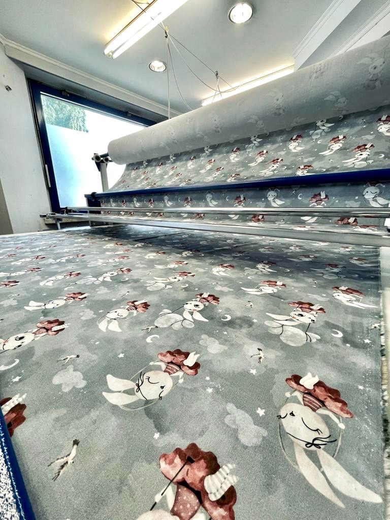 W Bytomiu, maszyna krojownicza rozkłada tkaninę z dziecięcym wzorem króliczków i chmurek, gotową do cięcia.