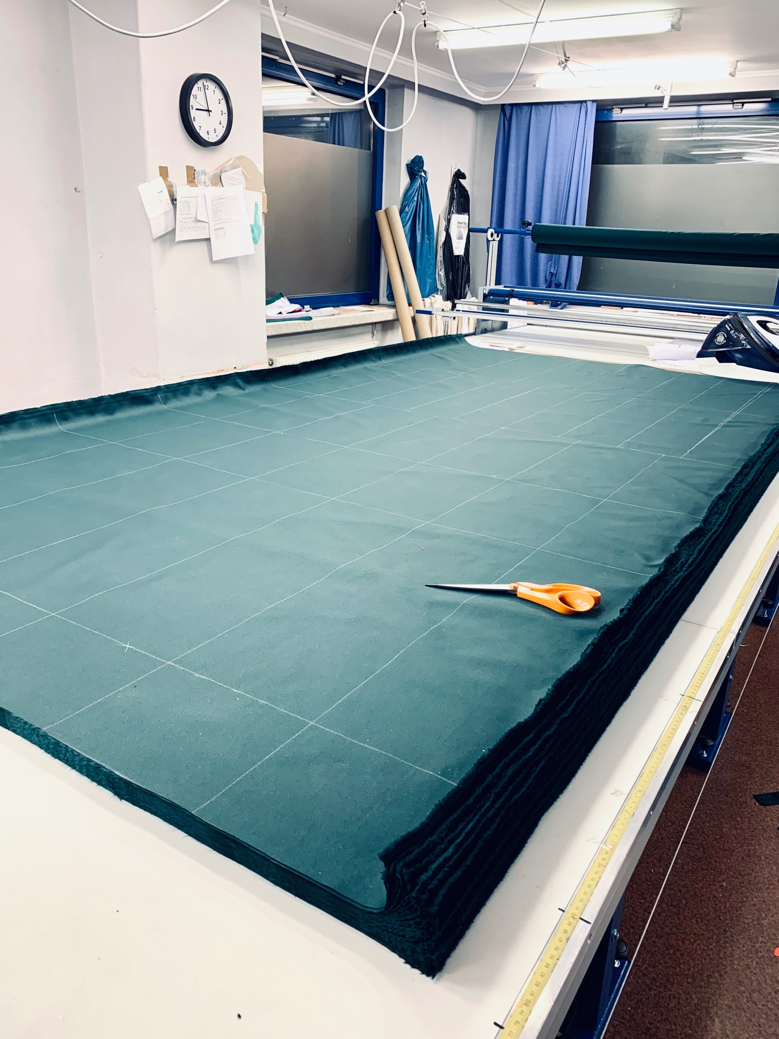 Stół krojczy z wieloma warstwami ciemnozielonego materiału w kratkę, nożyczki z pomarańczowymi uchwytami leżą na wierzchu, w tle widoczne wyposażenie krojowni.