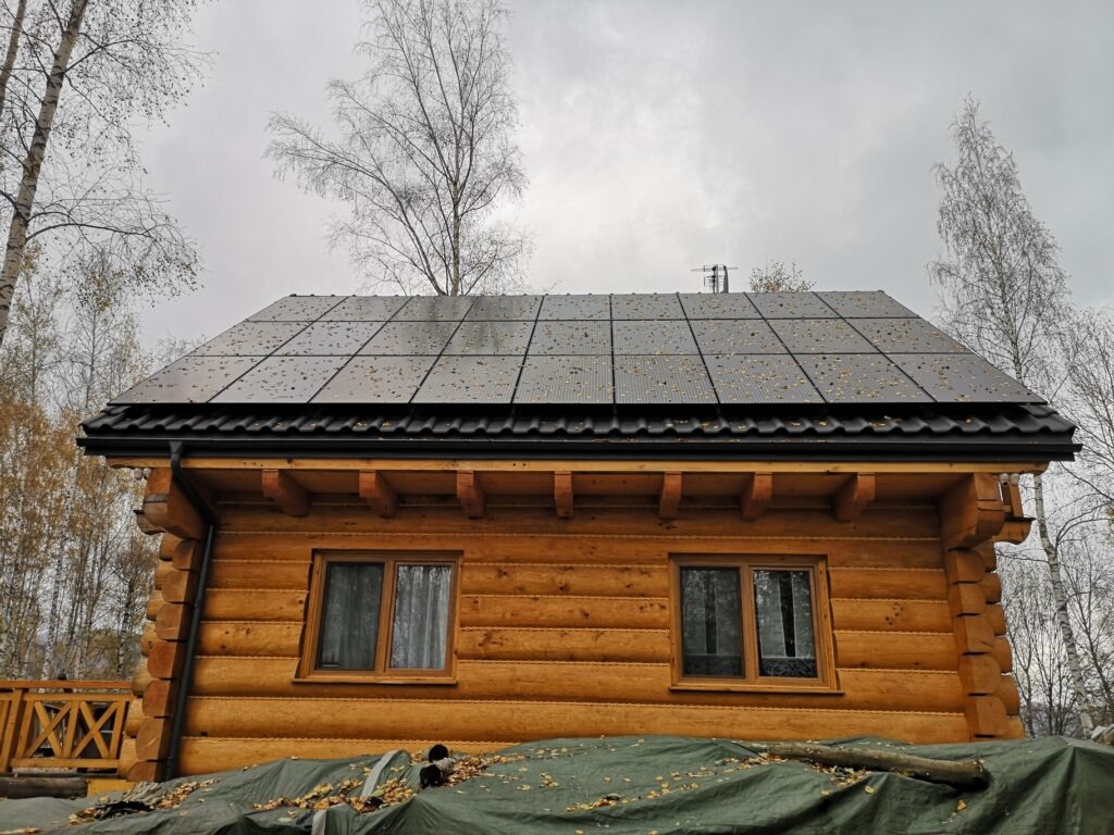 Panele słoneczne na dachu drewnianego domu z bali, pokryte opadłymi liśćmi, widoczne drzewa w tle i plandeka częściowo zakrywająca dół budynku.