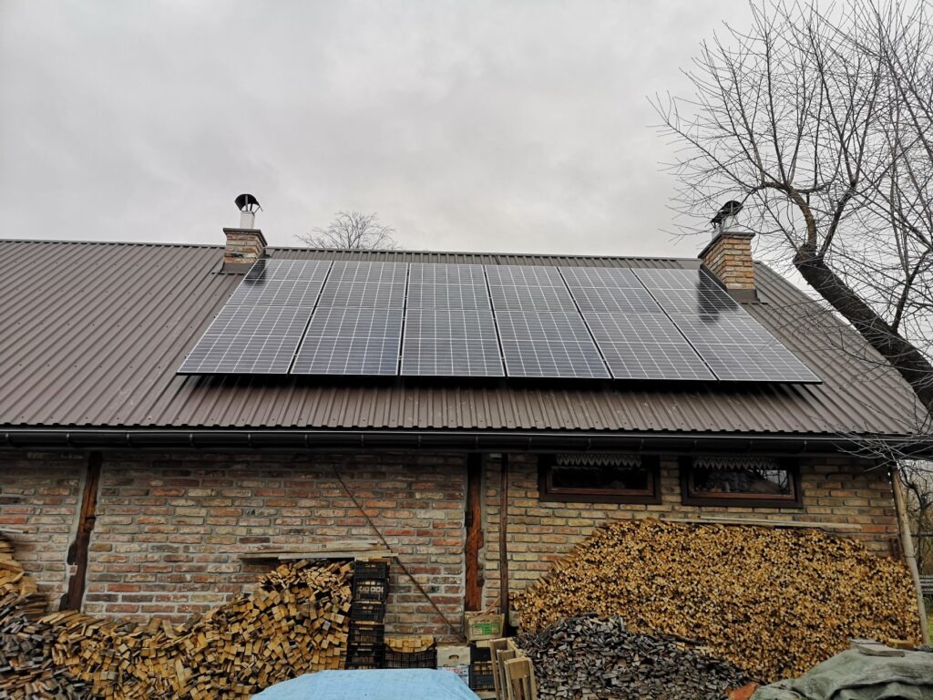 Panele słoneczne na brązowym, falistym dachu ceglanego budynku z dwoma kominami i stosem drewna opałowego z przodu, pochmurny dzień.