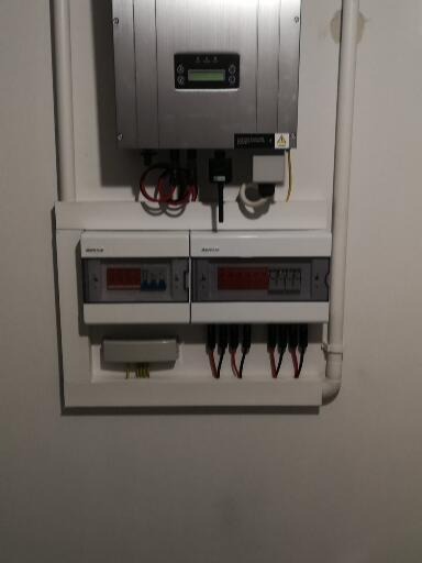 Srebrny inwerter fotowoltaiczny zamontowany na ścianie, poniżej skrzynka z bezpiecznikami i okablowaniem.