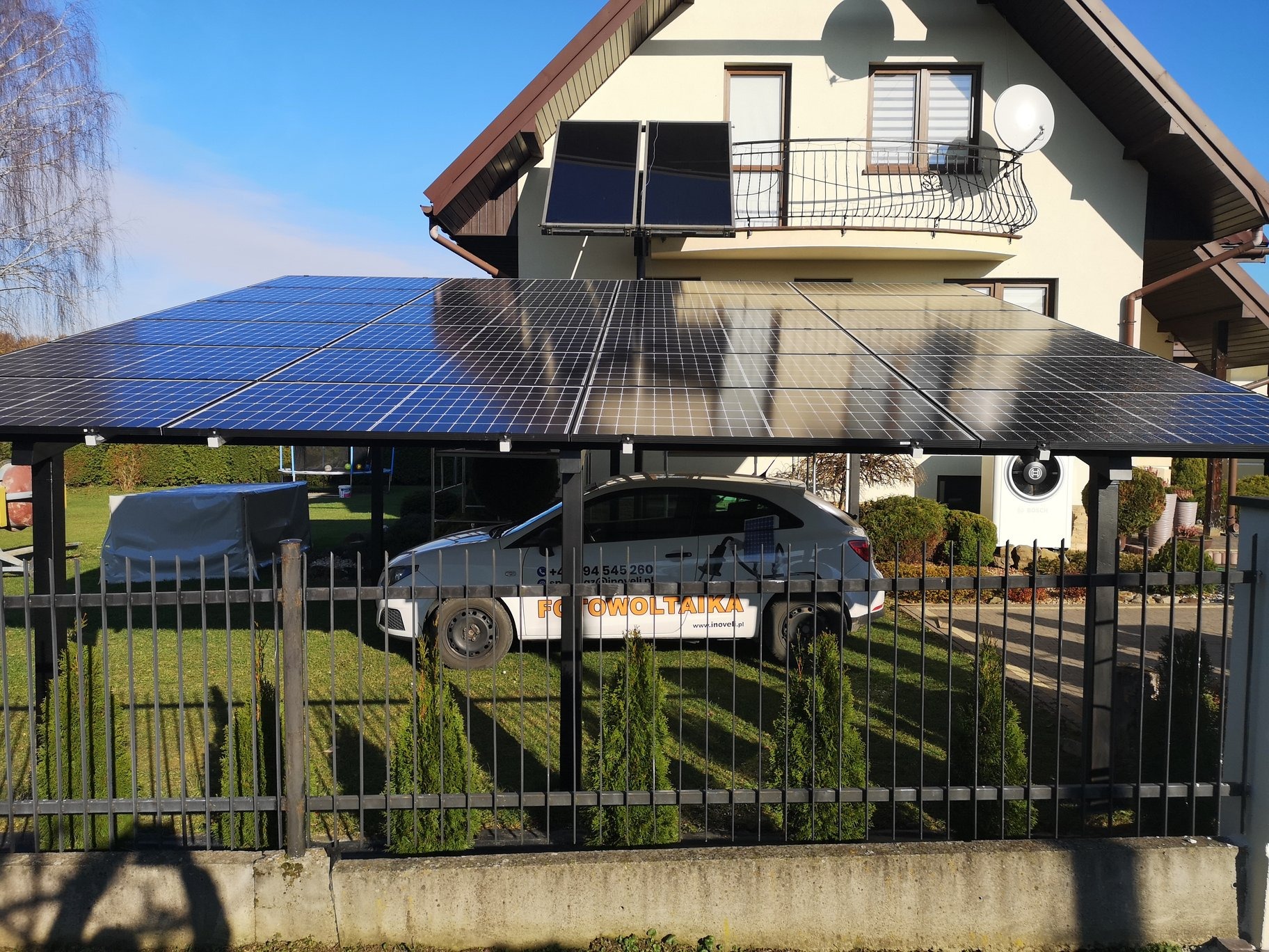 Instalacja fotowoltaiczna nad miejscem parkingowym dla samochodu, z dodatkowymi panelami na dachu domu jednorodzinnego w tle, widoczne logo firmy na pojeździe.