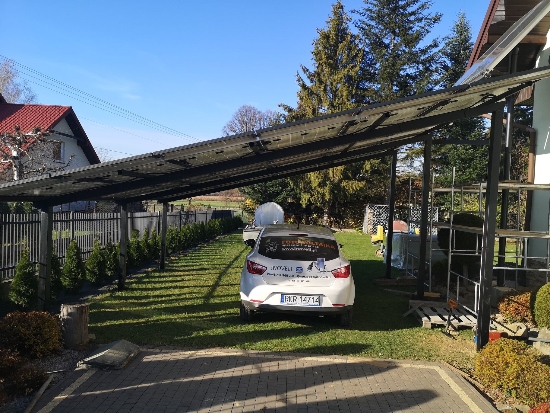 Zadaszenie fotowoltaiczne nad miejscem parkingowym, biały samochód z naklejką reklamową firmy fotowoltaicznej, widok z tyłu, trawnik, dom w tle.