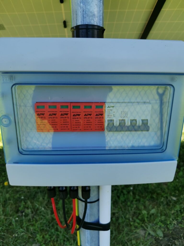 Skrzynka zabezpieczeń z ochronnikami przepięć APV OPS-3P-T2 w instalacji fotowoltaicznej, widoczne przewody i fragment panelu słonecznego.