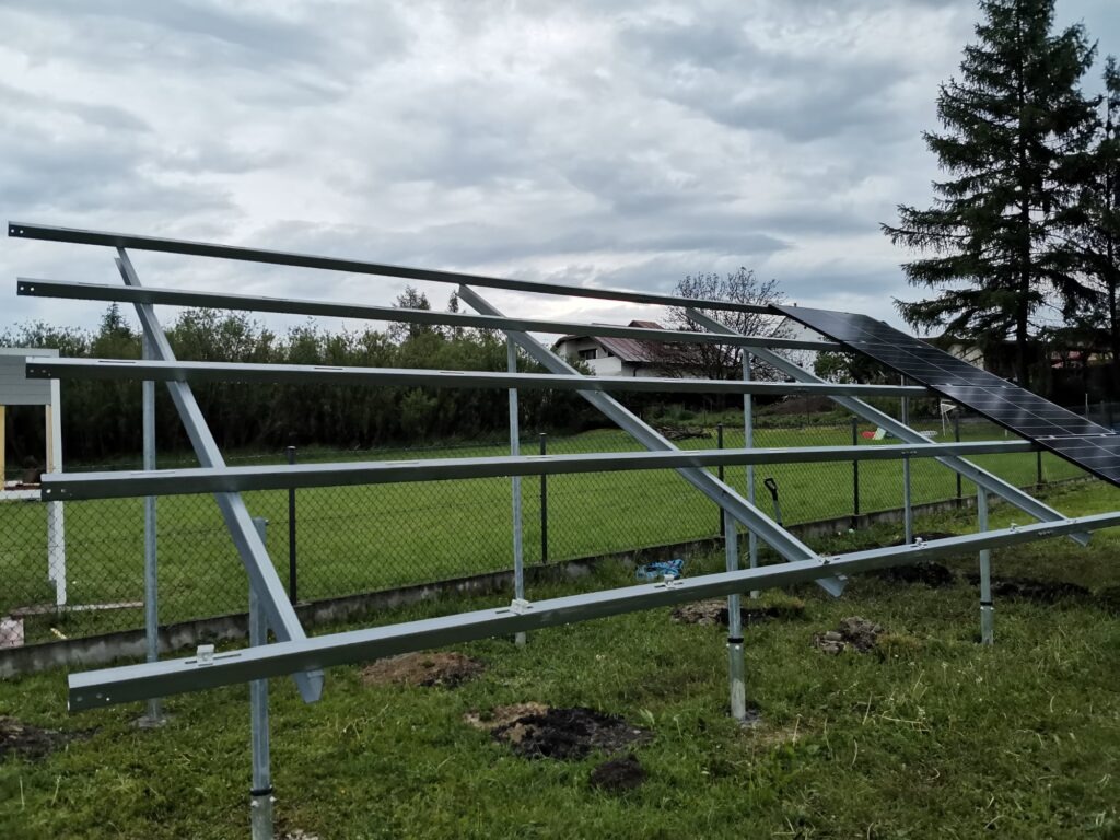 Stalowa konstrukcja wsporcza pod panele słoneczne na regulowanych słupkach, częściowo zamontowane panele, widok na ogród i dom w tle, pochmurne niebo.