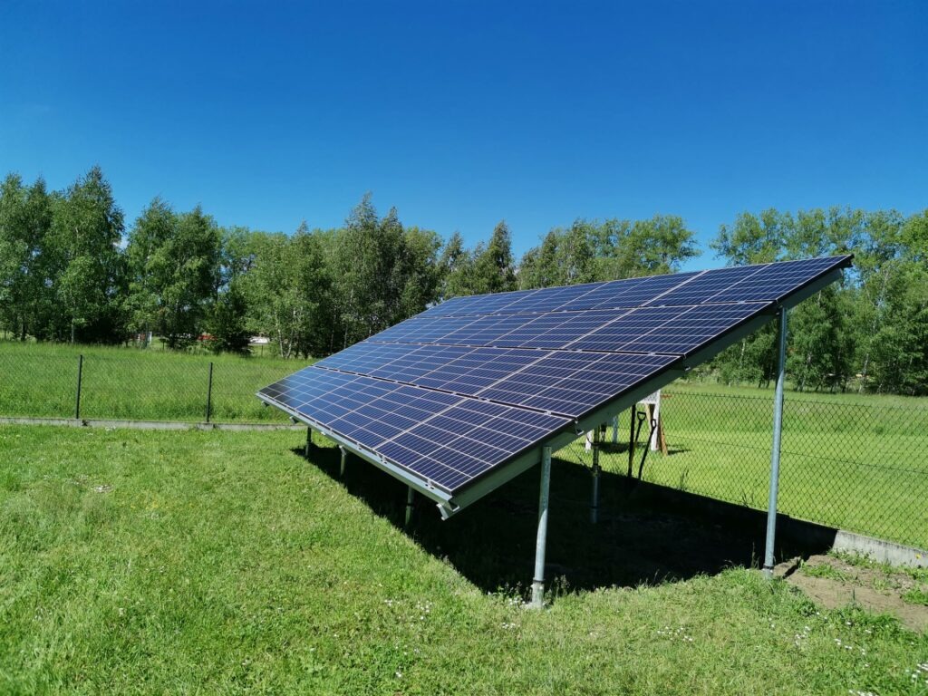 Panel słoneczny ustawiony na metalowej konstrukcji na trawniku, widok z boku, ogrodzenie z siatki w tle.