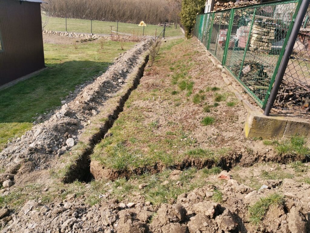 Wykopany rów w ziemi, prawdopodobnie pod kable do instalacji fotowoltaicznej, widoczny fragment ogrodu z zielonym ogrodzeniem i stertą drewna w tle.