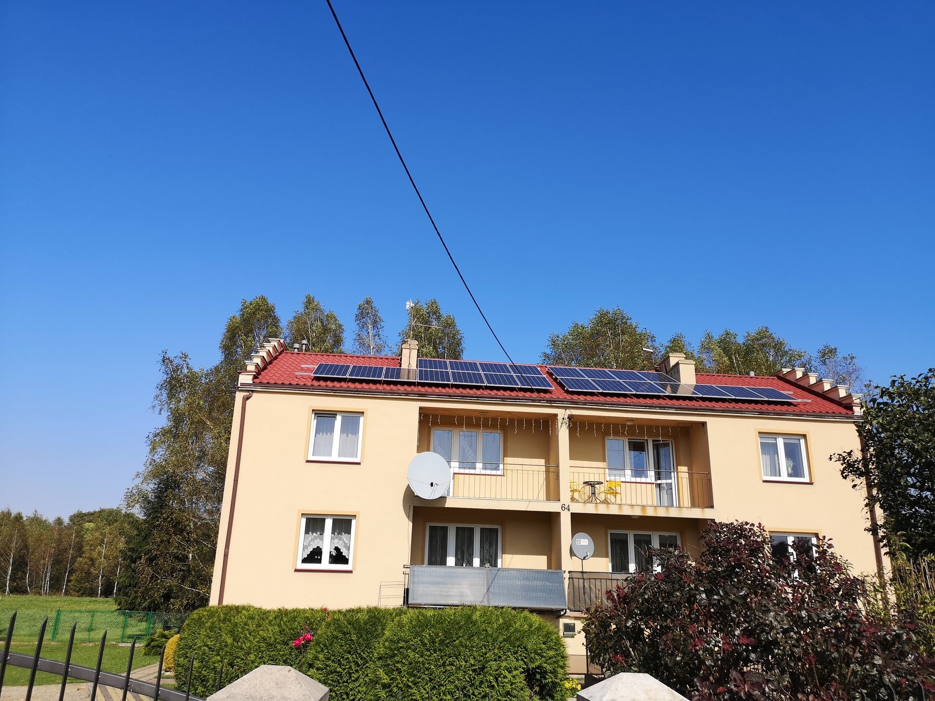 Żółty budynek mieszkalny z czerwonym dachem, na którym zamontowano panele słoneczne; widoczne anteny satelitarne na balkonach.