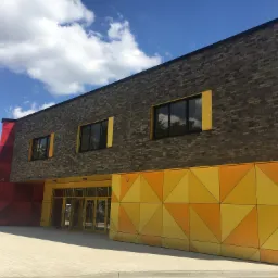 Nowoczesny budynek z cegły i panelami w geometryczne wzory w odcieniach żółci i czerwieni, przeszklone wejście z żółtymi ramami, okna z żółtymi obramowaniami, błękitne niebo z chmurami w tle.