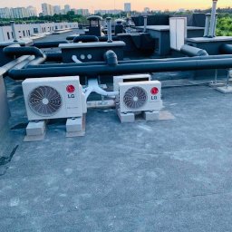 NOVA KLIMATYZACJE - Dwa agregaty klimatyzacyjne LG na dachu budynku z widokiem na panoramę miasta w tle, rury instalacyjne prowadzone po dachu.