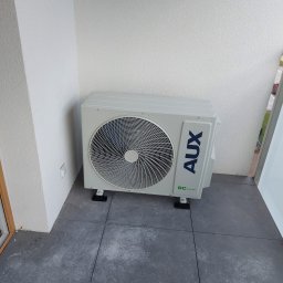 NOVA KLIMATYZACJE - Biała jednostka zewnętrzna klimatyzacji AUX, zamontowana na szarym balkonie, widok z przodu, z widocznym fragmentem ściany i okna. Minimalistyczny design.