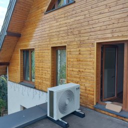 NOVA KLIMATYZACJE - Biała jednostka zewnętrzna klimatyzacji AUX zamontowana na dachu drewnianego domu z oknami i alarmem. Widok z dołu, lekki skos.