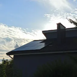 Panele słoneczne na ciemnoszarym dachu domu jednorodzinnego, widok z dołu na tle pochmurnego nieba z przebijającym słońcem.