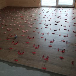 Klinkierek - Układanie płytek podłogowych imitujących drewno z użyciem czerwonych klipsów poziomujących i młotka w rogu pomieszczenia.