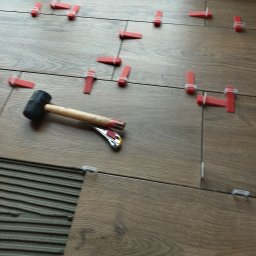 Klinkierek - Układanie płytek podłogowych imitujących drewno z użyciem systemu poziomowania płytek: czerwone kliny i klipsy, obok leży gumowy młotek.