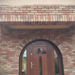 Klinkierek - Elewacja z cegły klinkierowej w Koszalinie, z łukowym wejściem i brązowymi drzwiami z szybkami, wykończona kolumnami z cegły.