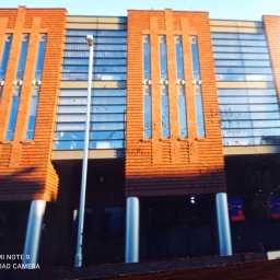 Klinkierek - Fragment elewacji budynku z cegły klinkierowej z pionowymi oknami, widoczne kolumny i lampa uliczna.