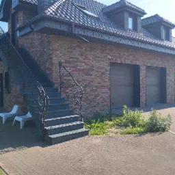 Dwukondygnacyjny dom z cegły klinkierowej z grafitową dachówką, ozdobnymi schodami zewnętrznymi z metalową balustradą i dwoma bramami garażowymi.