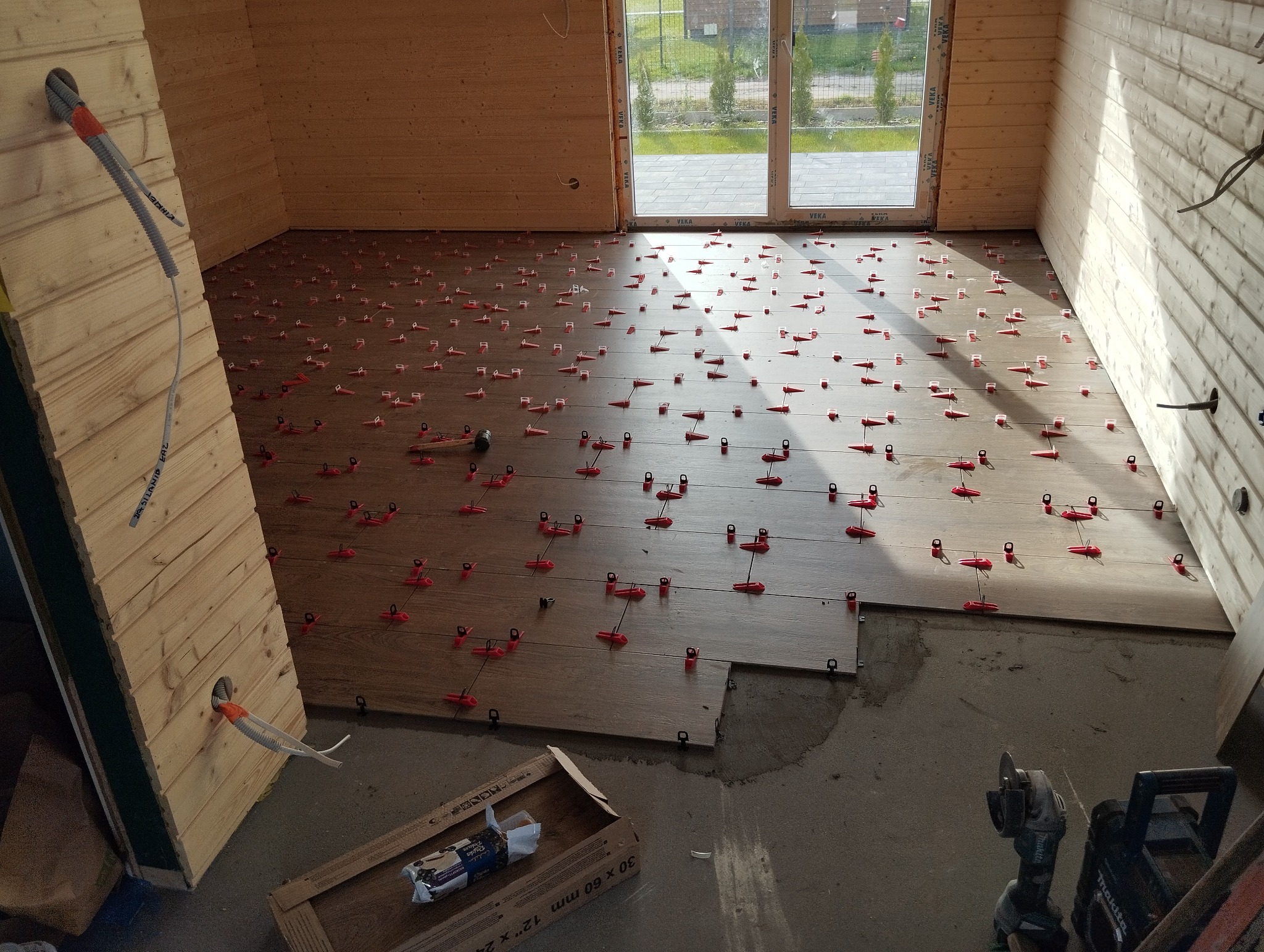 Układanie płytek podłogowych imitujących drewno w drewnianym domku, z użyciem systemu poziomowania płytek (czerwone klipsy i kliny), widoczne narzędzia glazurnicze i opakowanie płytek.