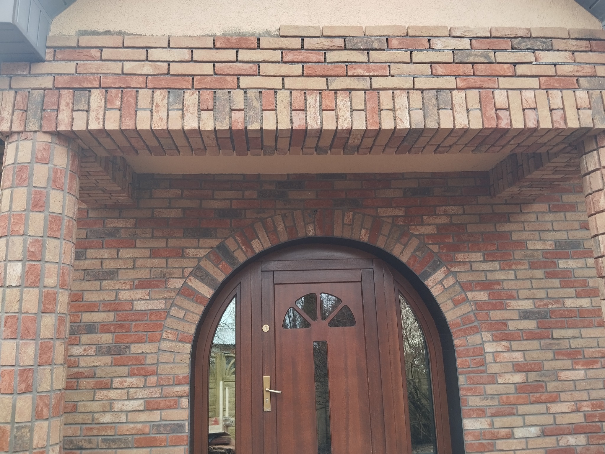 Elewacja z cegły klinkierowej w Koszalinie, z łukowym wejściem i brązowymi drzwiami z szybkami, wykończona kolumnami z cegły.