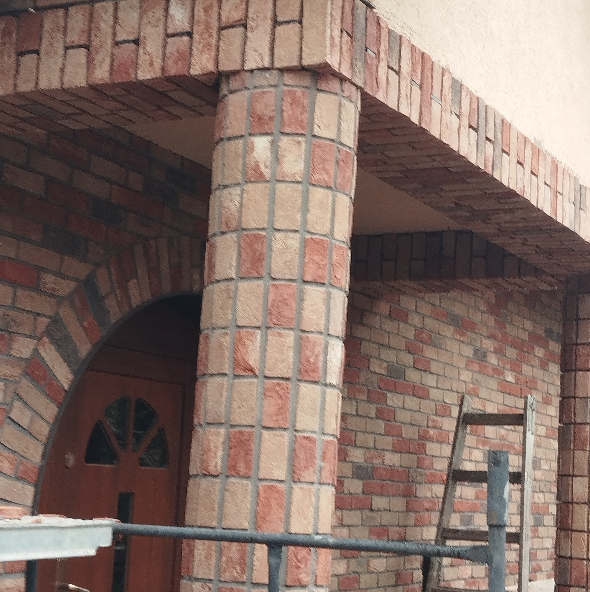 Ceglana kolumna i łuk nad drzwiami wejściowymi, z widoczną drabinką opartą o ścianę z cegły w tle, w trakcie budowy lub renowacji.