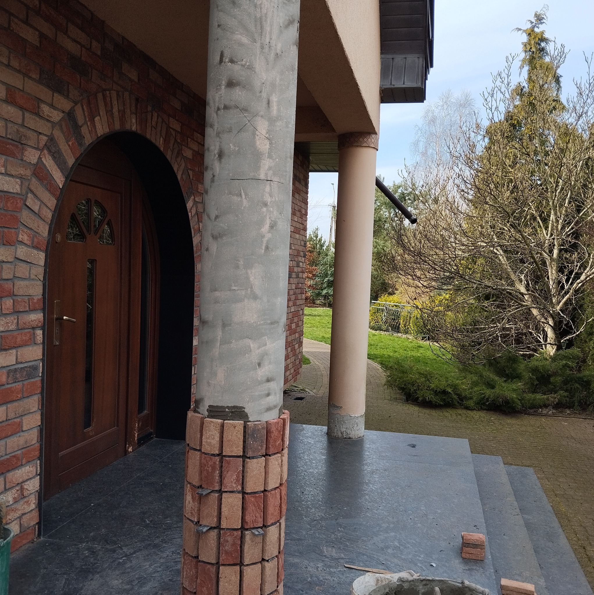 Murowana kolumna w trakcie budowy, z widocznym szarym betonem i fragmentami cegieł, przylegająca do ceglanego łuku nad drzwiami wejściowymi, na tle ogrodu.