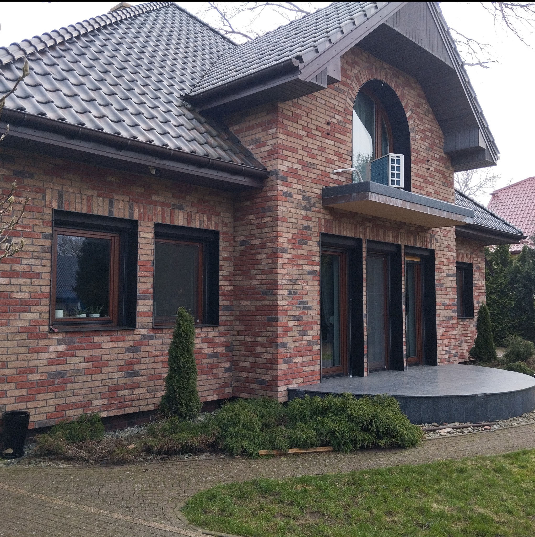 Dwukondygnacyjny dom z cegły klinkierowej z ciemną dachówką, okna z ciemnymi ramami, zaokrąglony taras z ciemnej płyty, małe drzewka iglaste w ogrodzie.