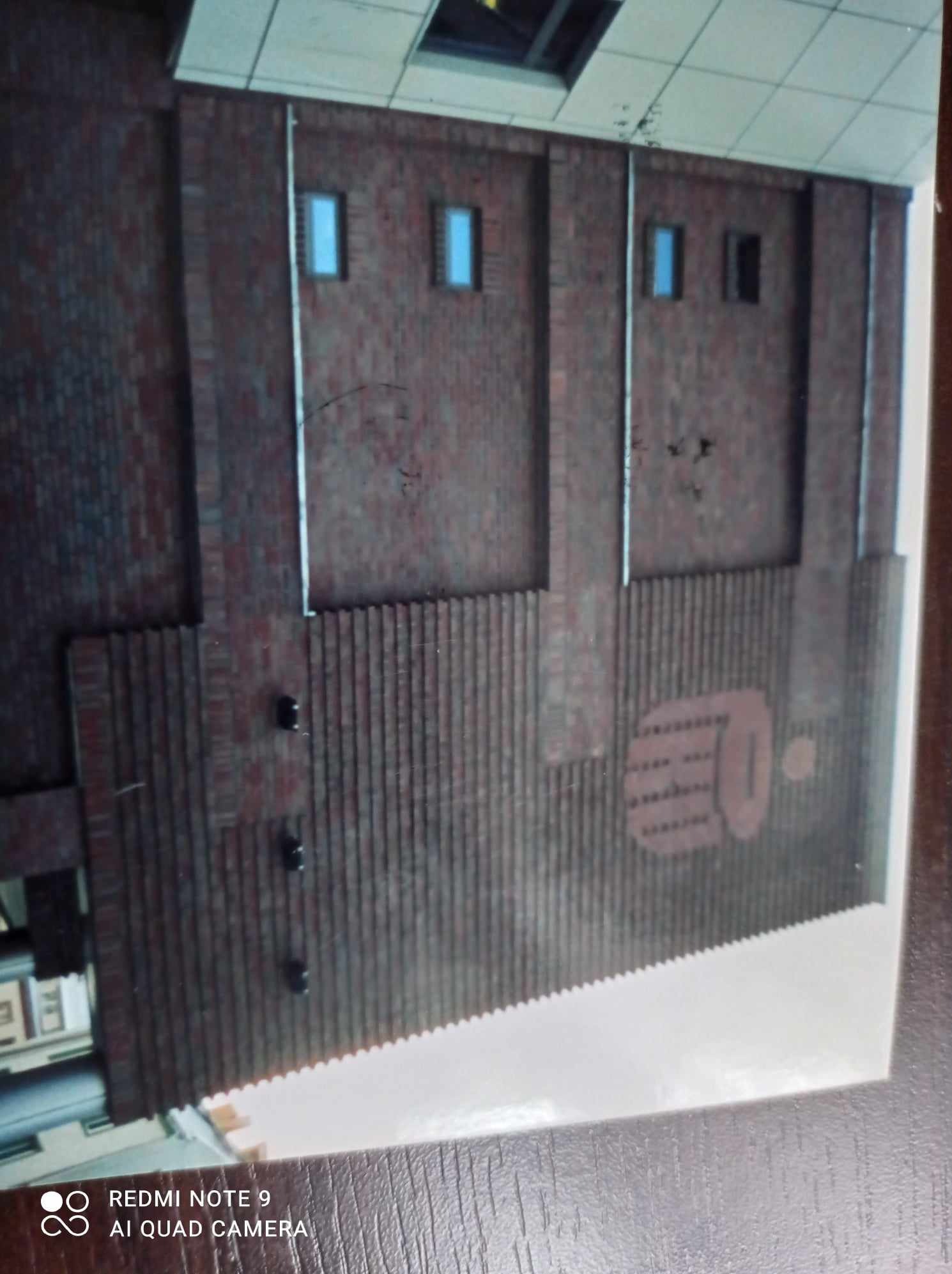 Ceglana elewacja budynku z małymi oknami i dekoracyjnym elementem z pionowych żeberek oraz wzorem graficznym w kolorze cegły, sfotografowana pod kątem.