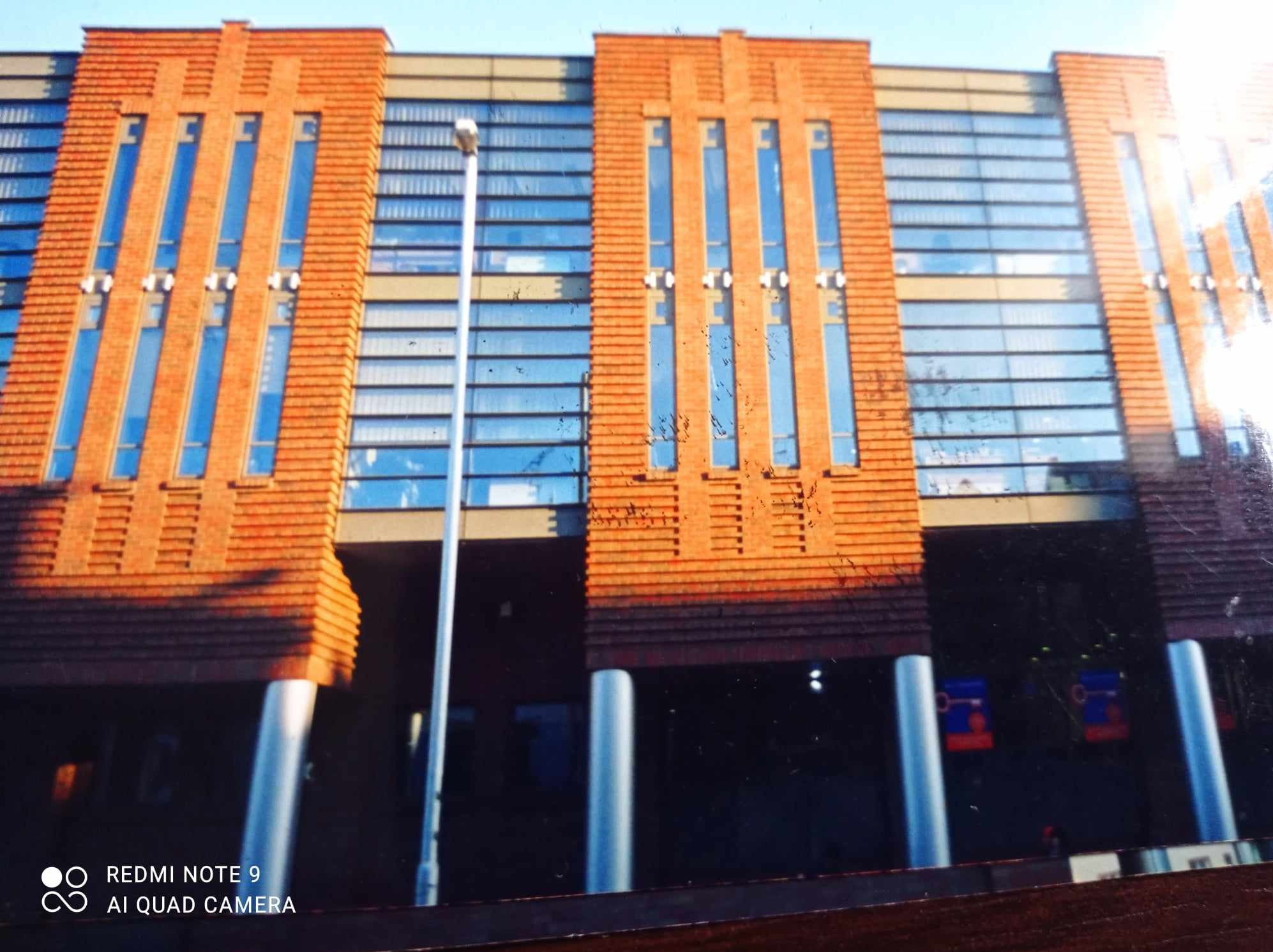 Fragment elewacji budynku z cegły klinkierowej z pionowymi oknami, widoczne kolumny i lampa uliczna.