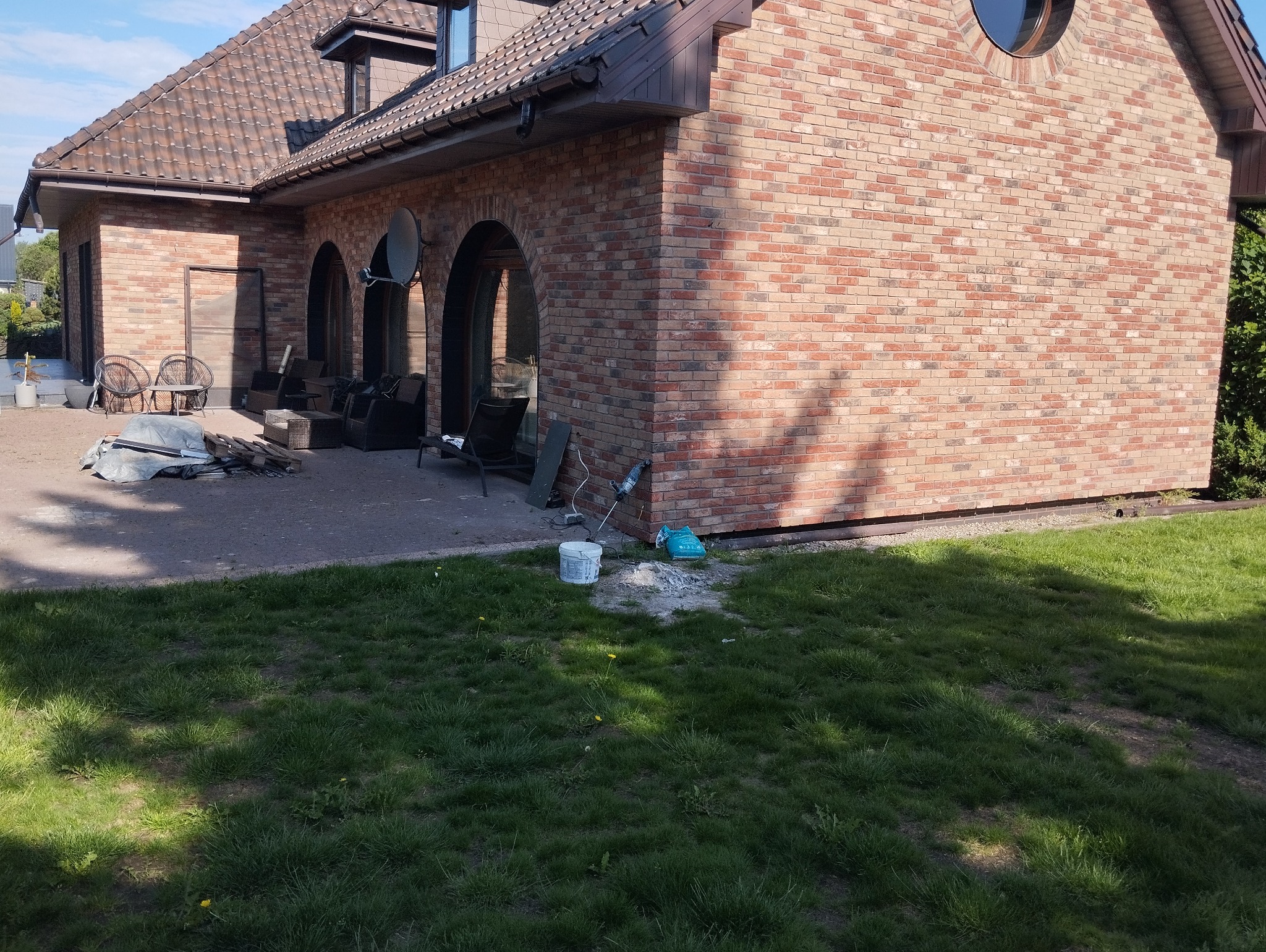Murowany dom z cegły klinkierowej w trakcie budowy, widoczne narzędzia i materiały budowlane na zewnątrz, trawnik przed budynkiem.