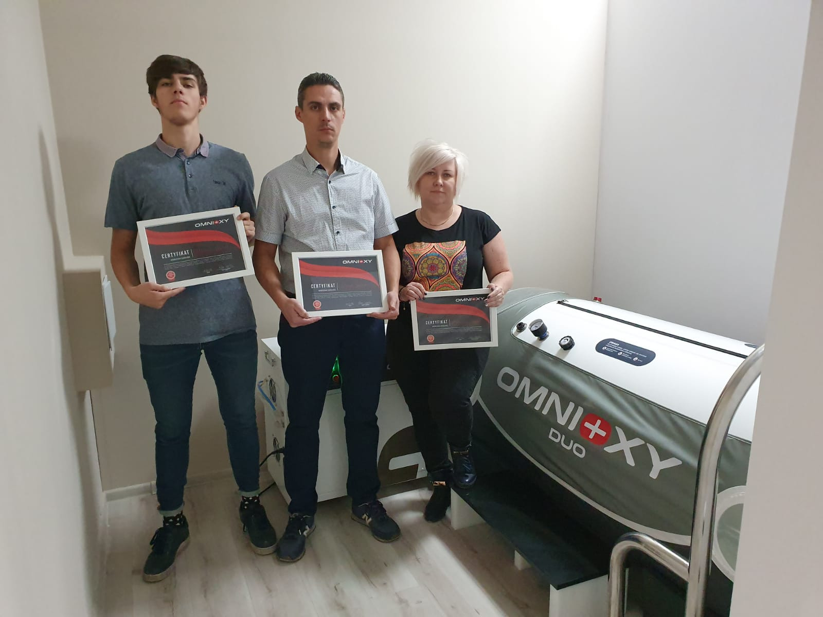 Trzy osoby trzymają certyfikaty OMNI+XY obok komory hiperbarycznej OMNI+XY DUO w jasnym pomieszczeniu.