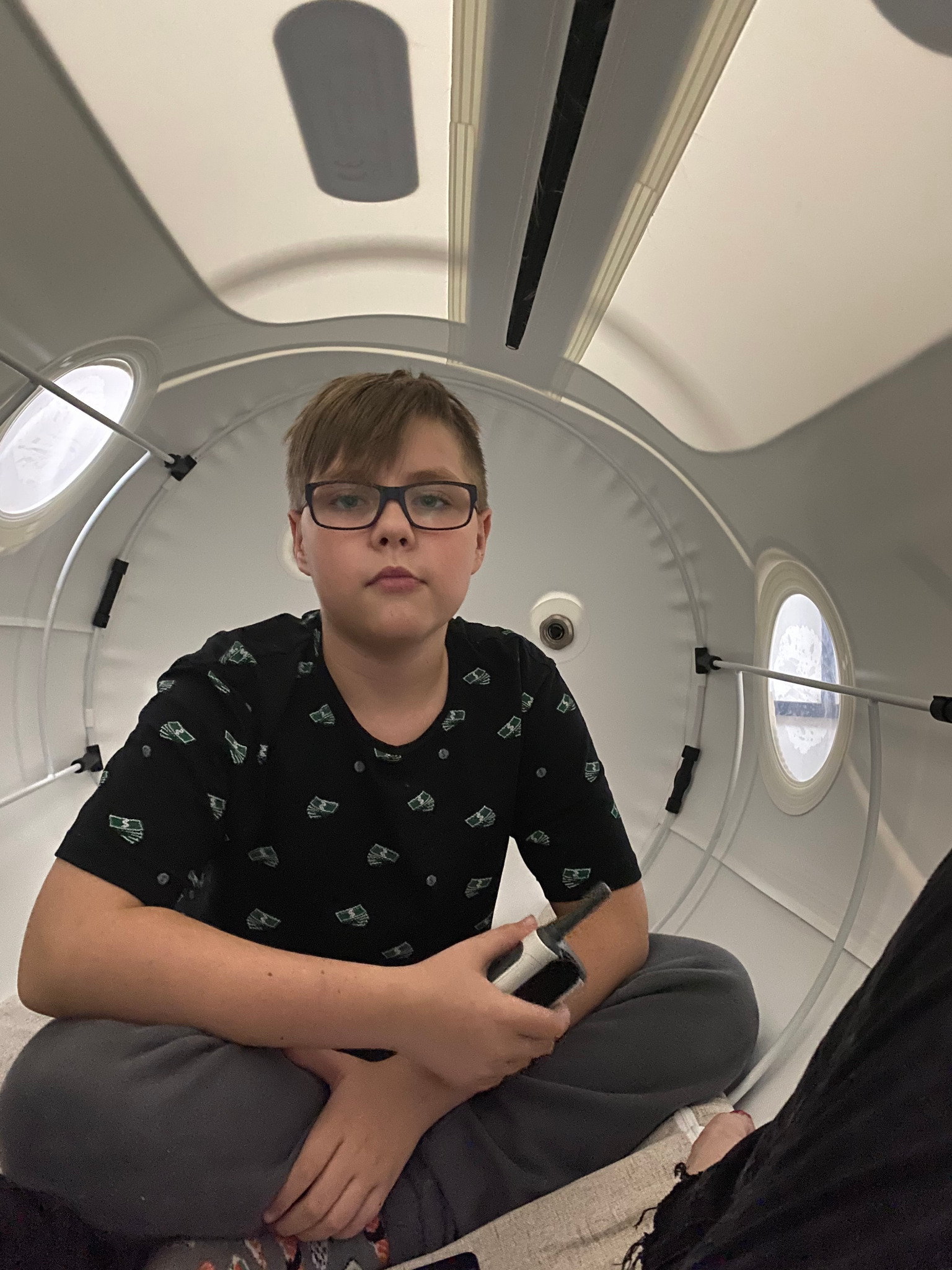 Młody mężczyzna w okularach siedzi wewnątrz hiperbarycznej komory tlenowej, trzymając w dłoni małe urządzenie elektroniczne.