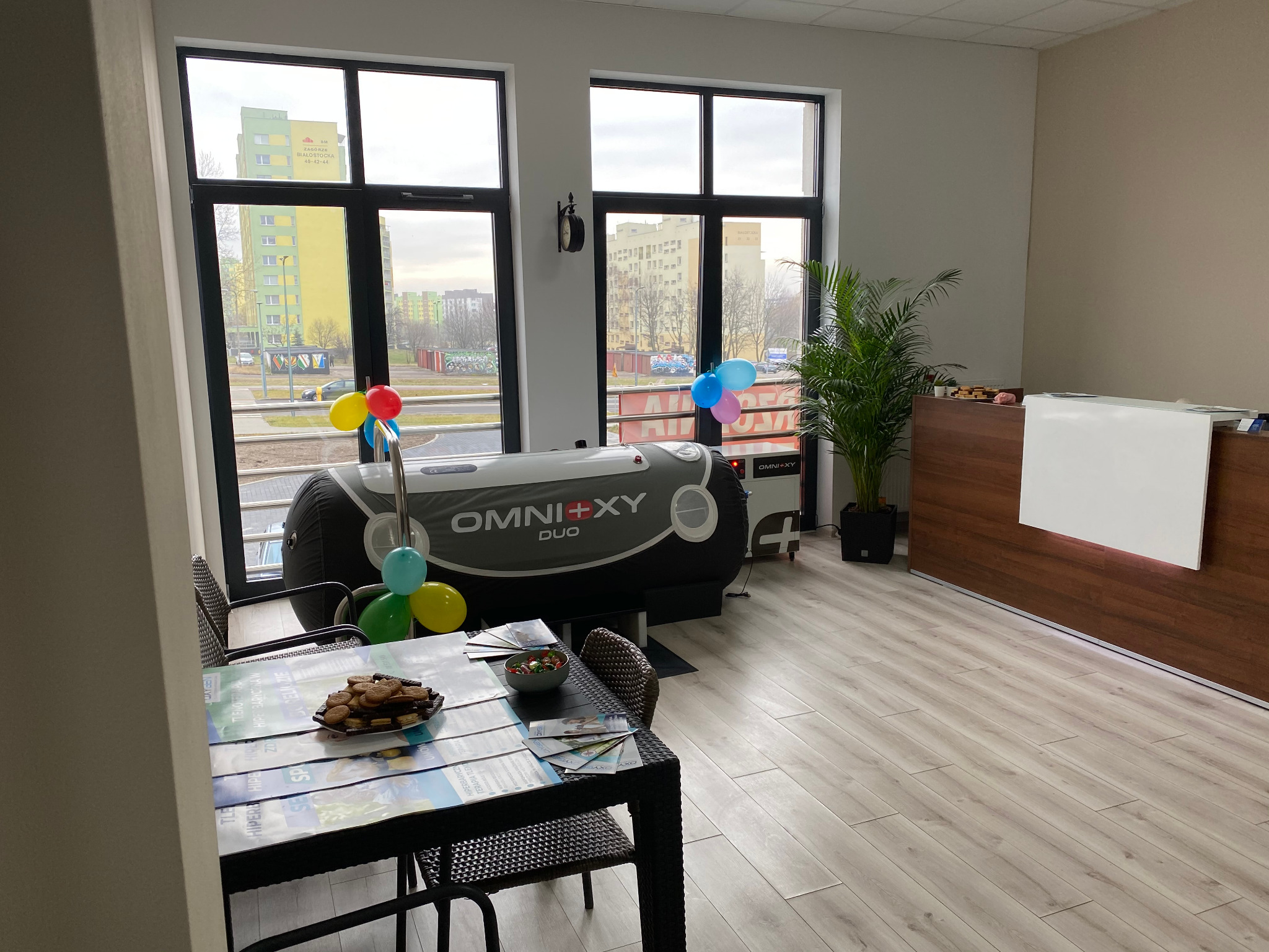 Wnętrze gabinetu fizjoterapii z komorą hiperbaryczną Omni+XY Duo udekorowaną balonami, stół z poczęstunkiem i ulotkami informacyjnymi dla klientów.