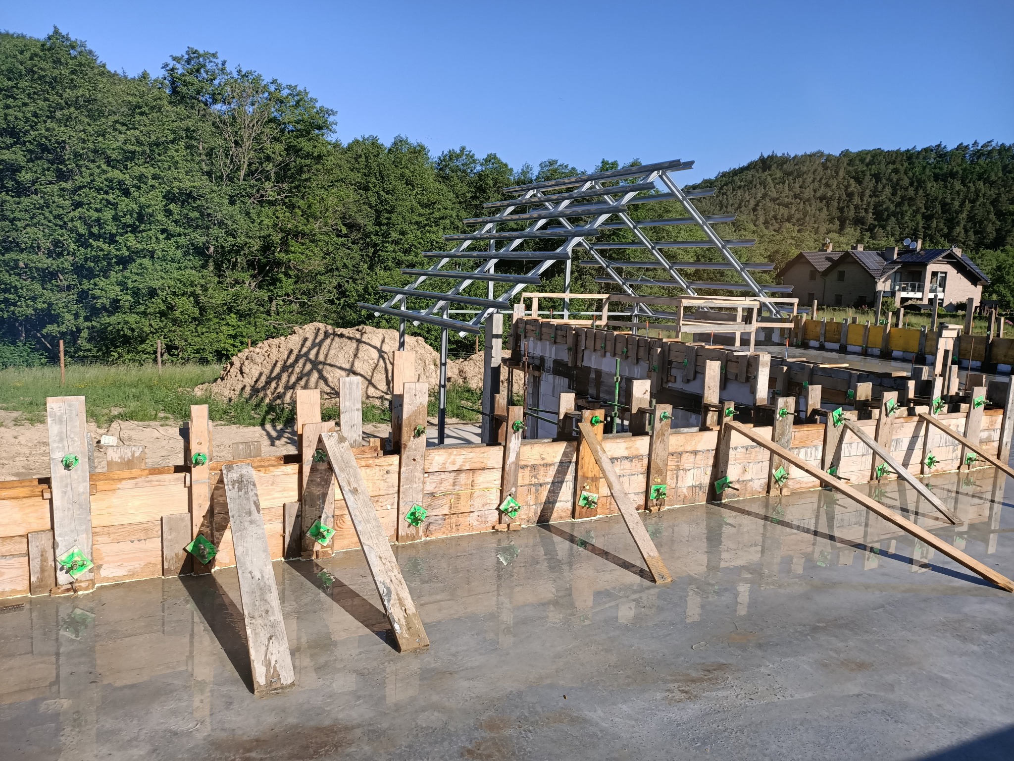 Wylany fundament z drewnianym szalunkiem wzmocnionym ukośnymi belkami i zielonymi łącznikami; w tle konstrukcja dachu z metalowych profili, pagórkowaty teren z domami i lasem w słoneczny dzień.